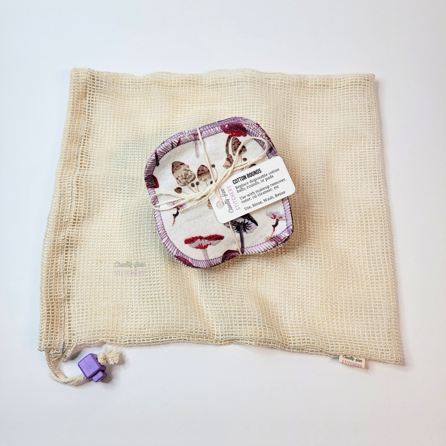 A bundled pack of the mushrooms print cotton rounds laid on top of an organic cotton mesh washing bag. It is an off white natural color with square mesh, and a lilac purple cord stop on the drawstring.