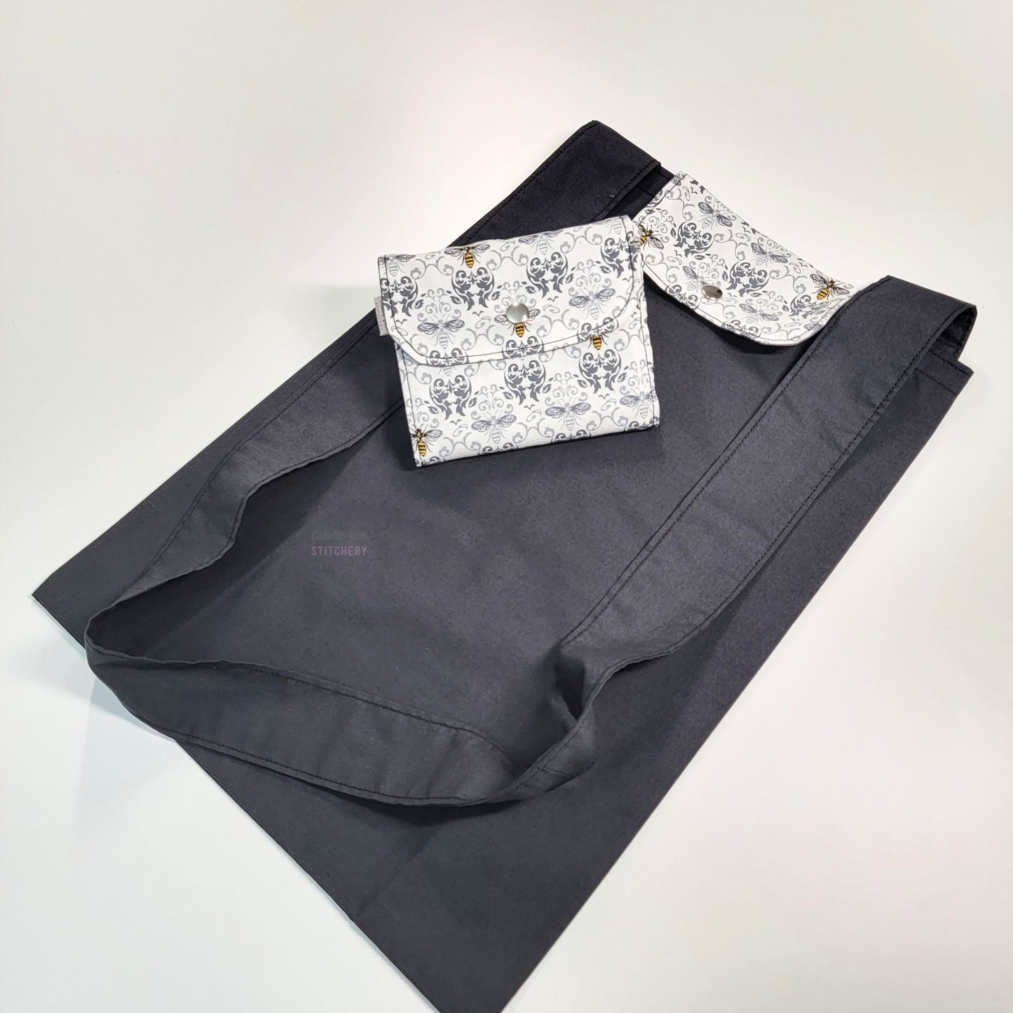 A black tote bag with a long strap and a flap with a white and grey damask and bees print. On top of the bag is another bag that has been folded up and snapped together like a wallet.