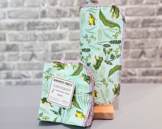 Blue Frogs NonPaper Towels - cute little blushing green and yellow frogs with lily pads, lotus flowers, mushrooms, cattails, and other plants on a light aqua blue background. Shown here on a wooden NonPaper Towel holder next to a bundled pack.
