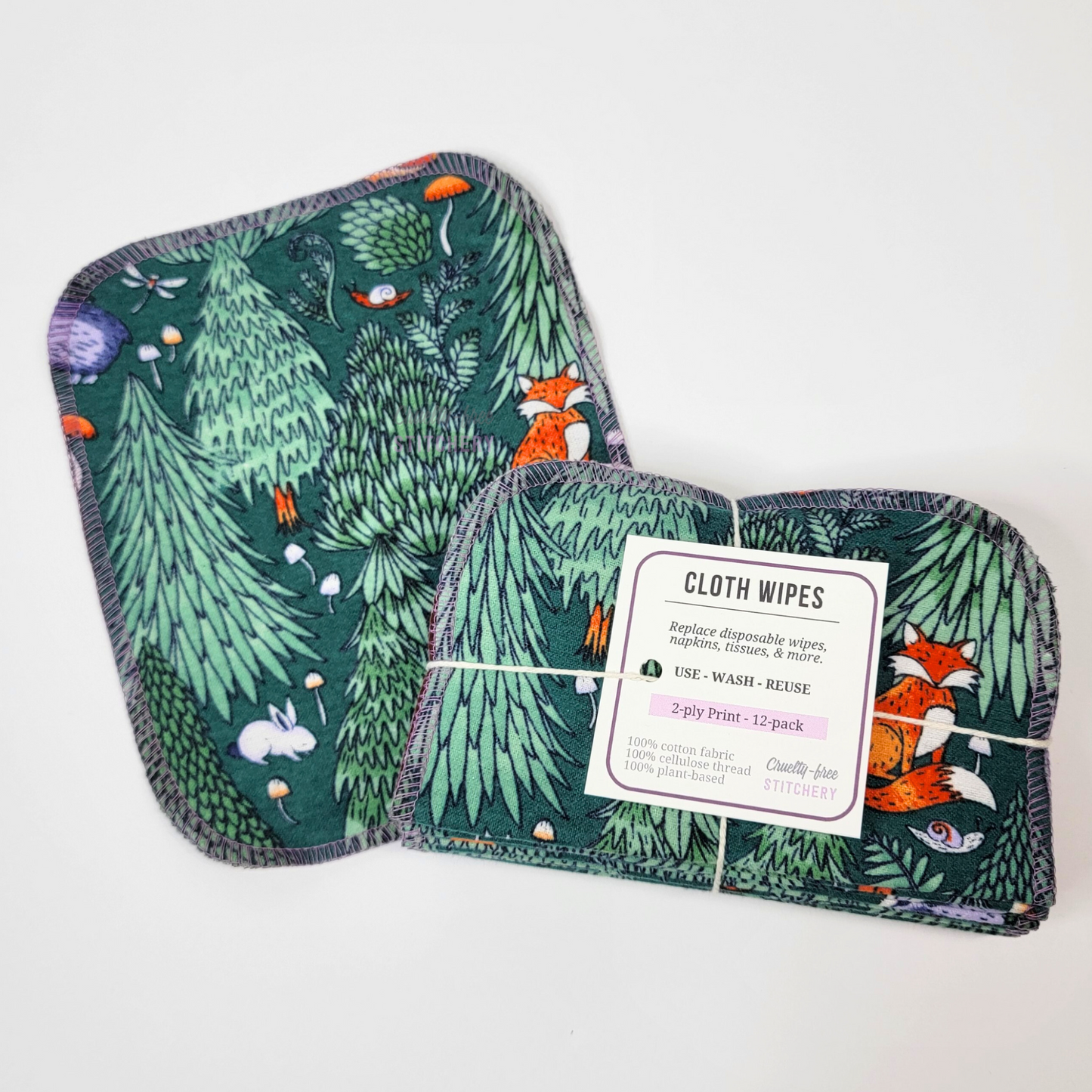 A woodland cloth wipe next to a bundled pack. The wipes are a rounded rectangle shape, stitched around the edges with a lilac purple thread. The bundle is tied with a string like a gift, and has a tag.