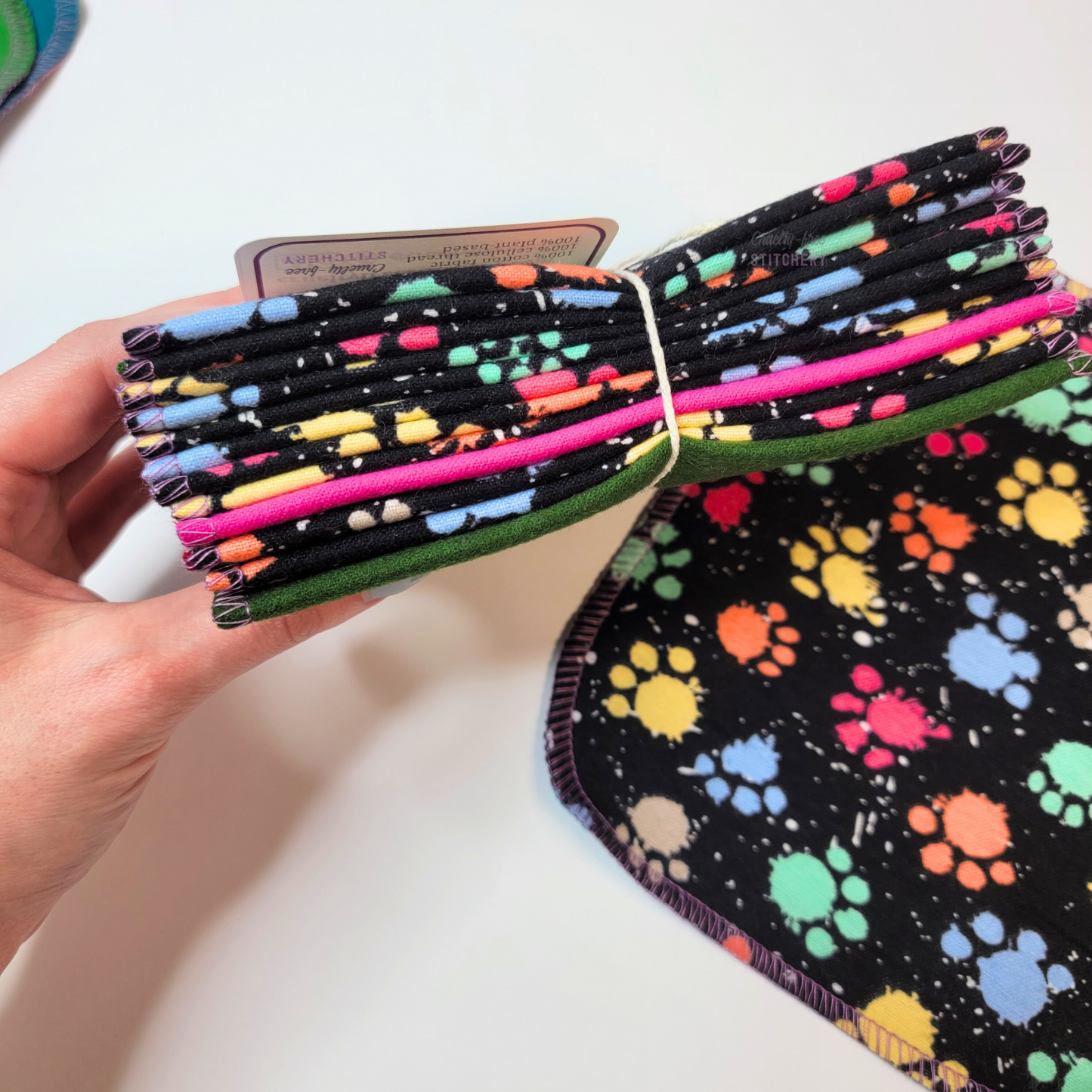 A bundle of the rainbow paw print paint splatter cloth wipes in my hand, pictured from the bottom to show there are twelve wipes in the pack. Two random wipes in the bundle are folded opposite, to show the different colors.