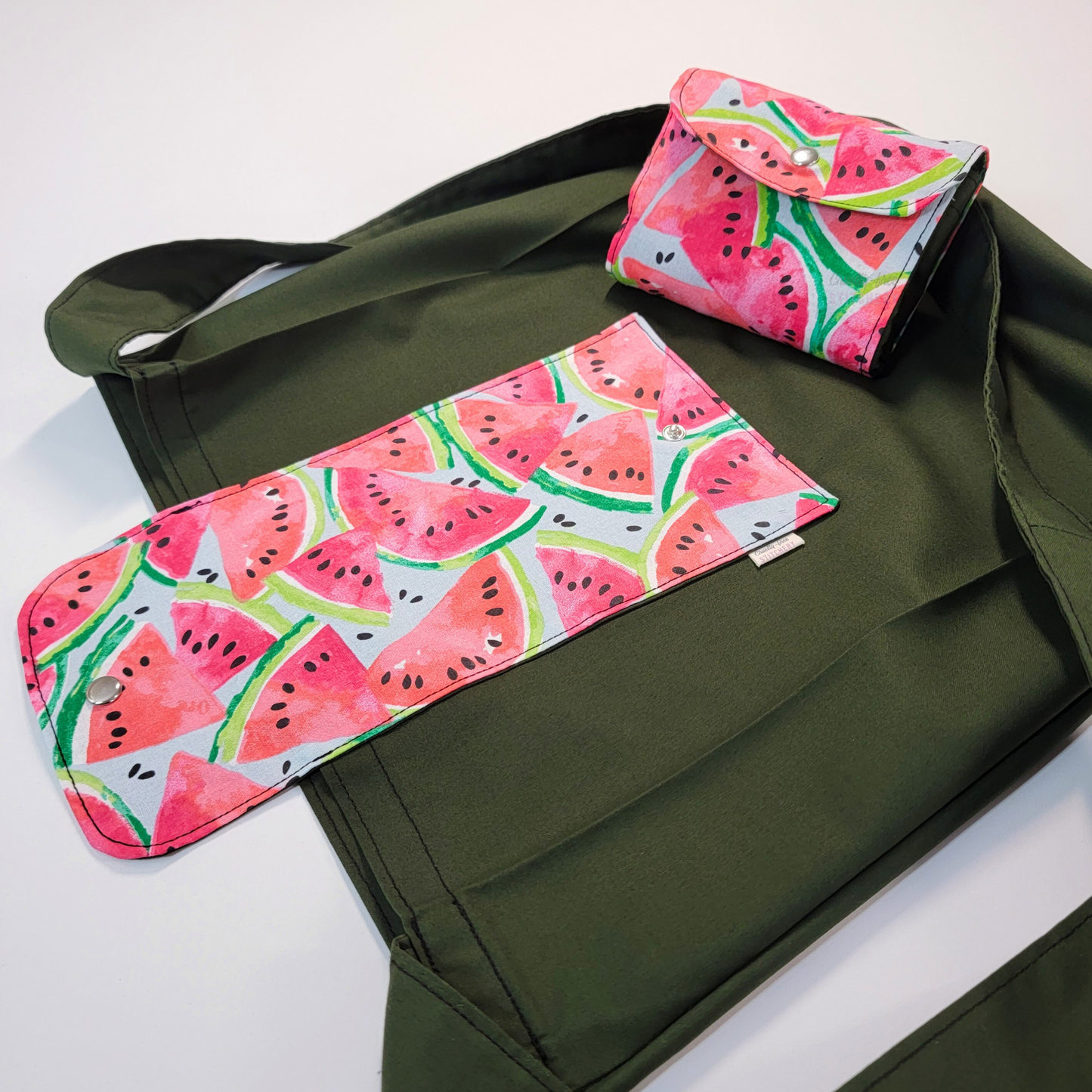 A solid dark forest green tote bag with a long strap and a light blue flap printed with watermelon slices in a painterly style. On top of the bag is another bag that has been folded up and snapped together like a wallet.