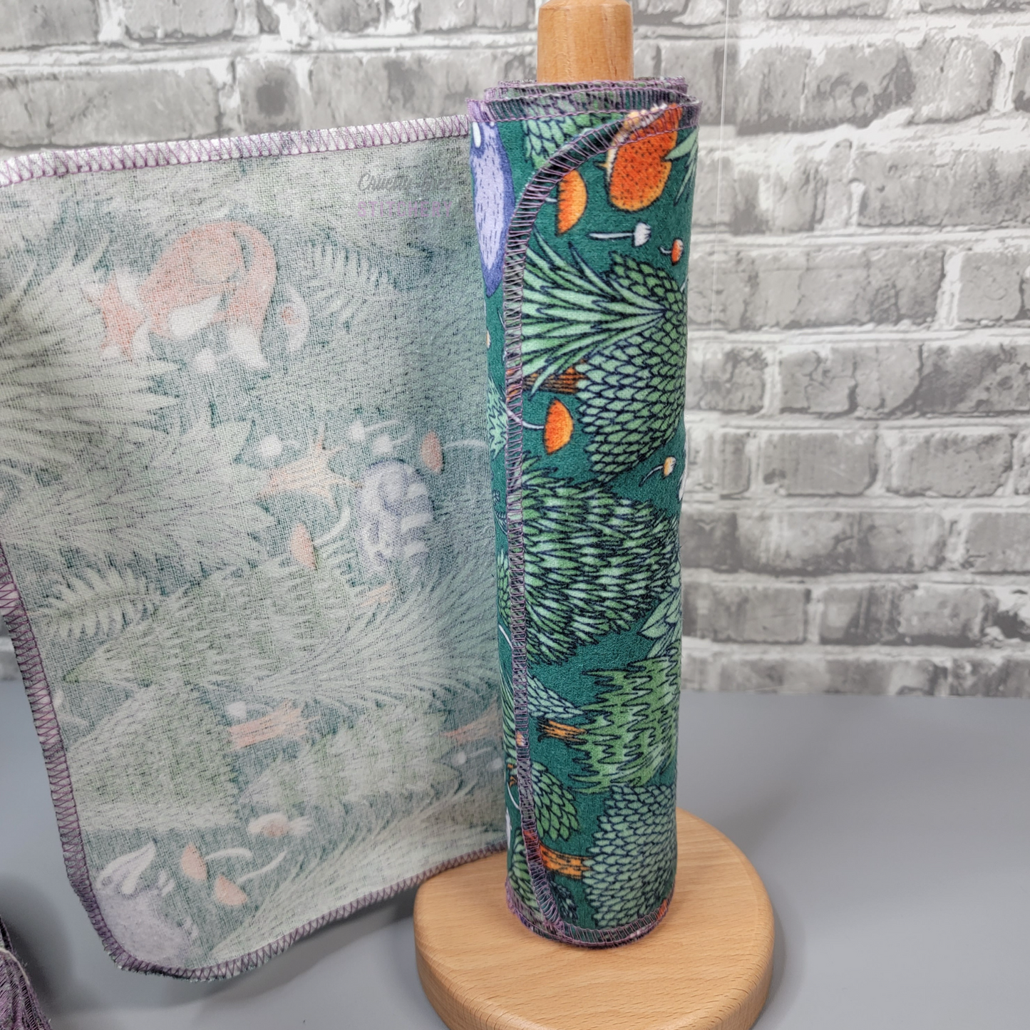 A woodland print NonPaper towel being pulled off the roll, showing they are a single layer of fabric. Stitched around the edges with a lilac purple thread.