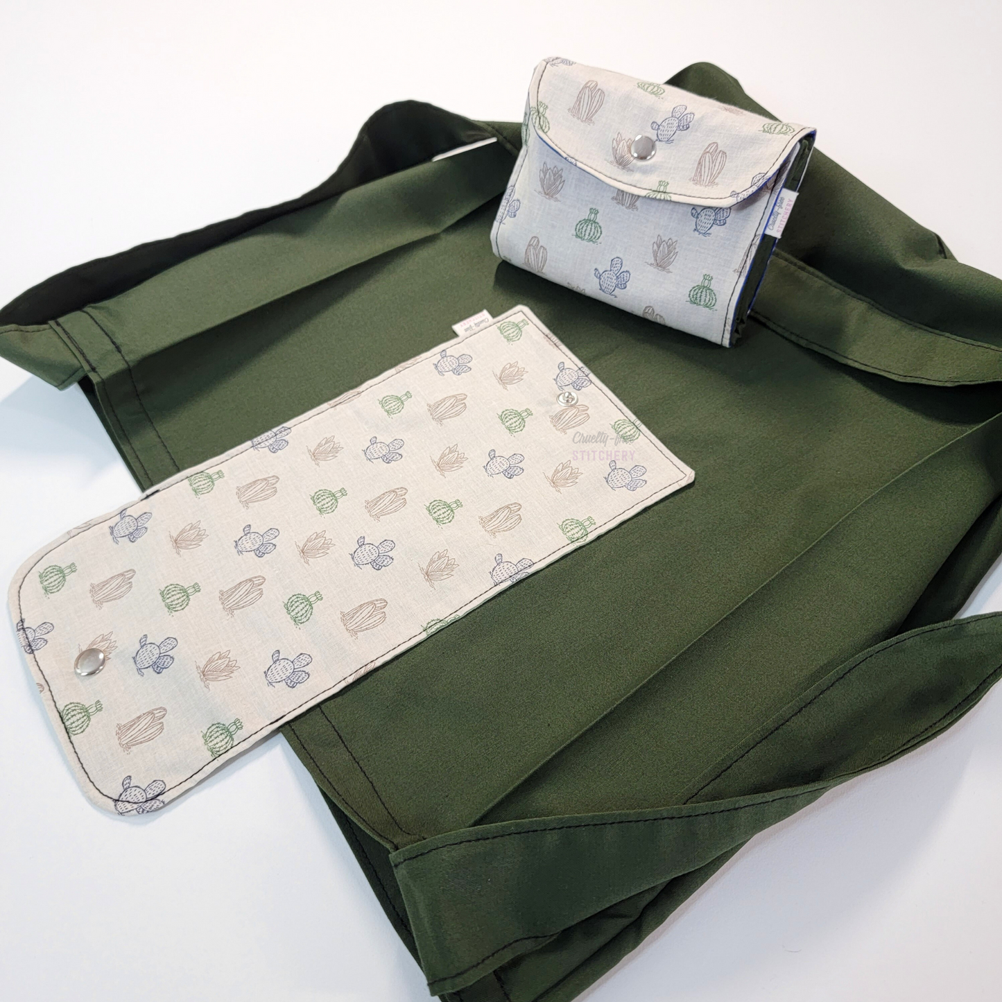 A solid dark forest green tote bag with a long strap and a light tan flap printed with small cactus icons in brown, green, and blue. On top of the bag is another bag that has been folded up and snapped together like a wallet.