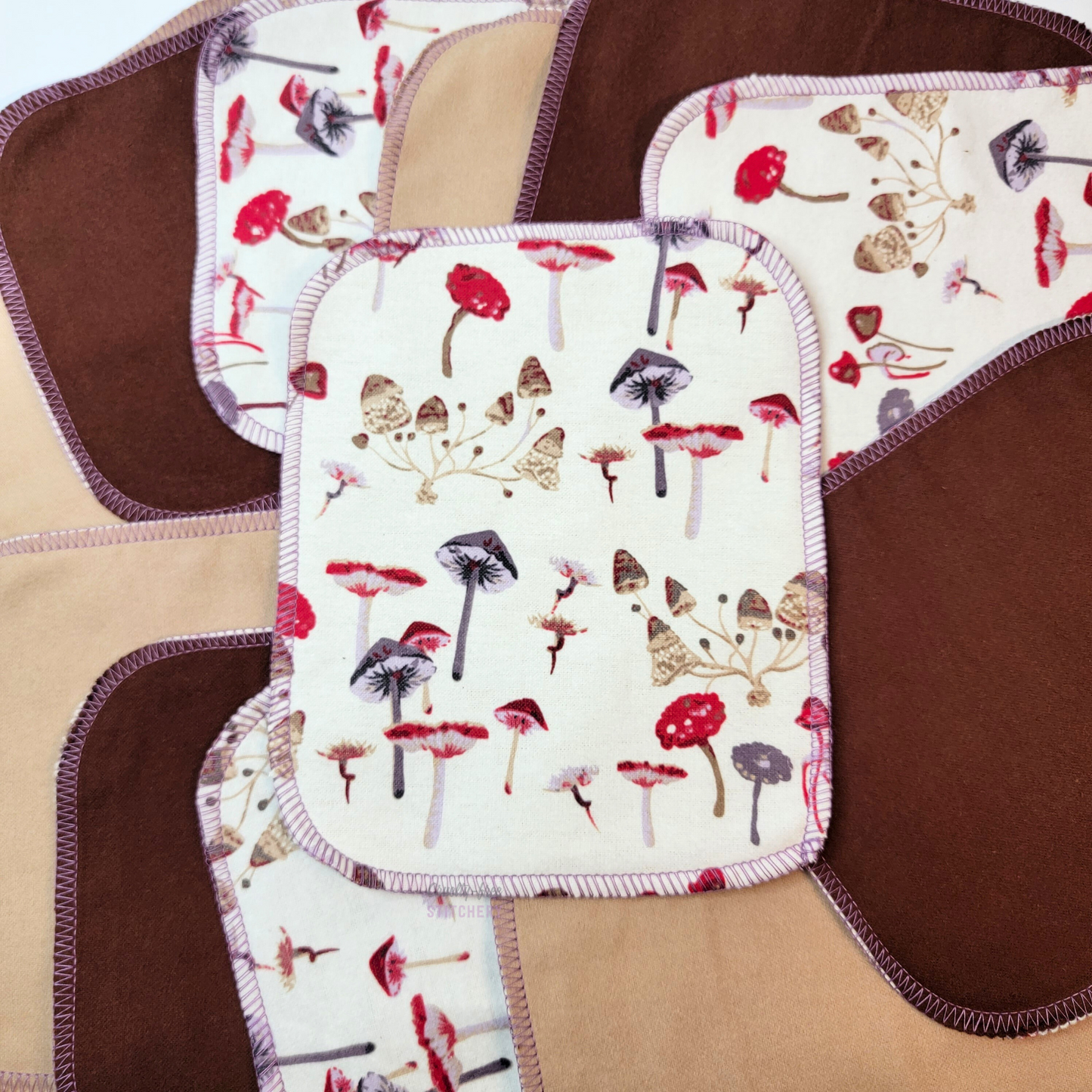 Several mushrooms print cloth wipes scattered on the table, some upside down to show the brown shades on the back, and one in the center showing the full mushrooms print.