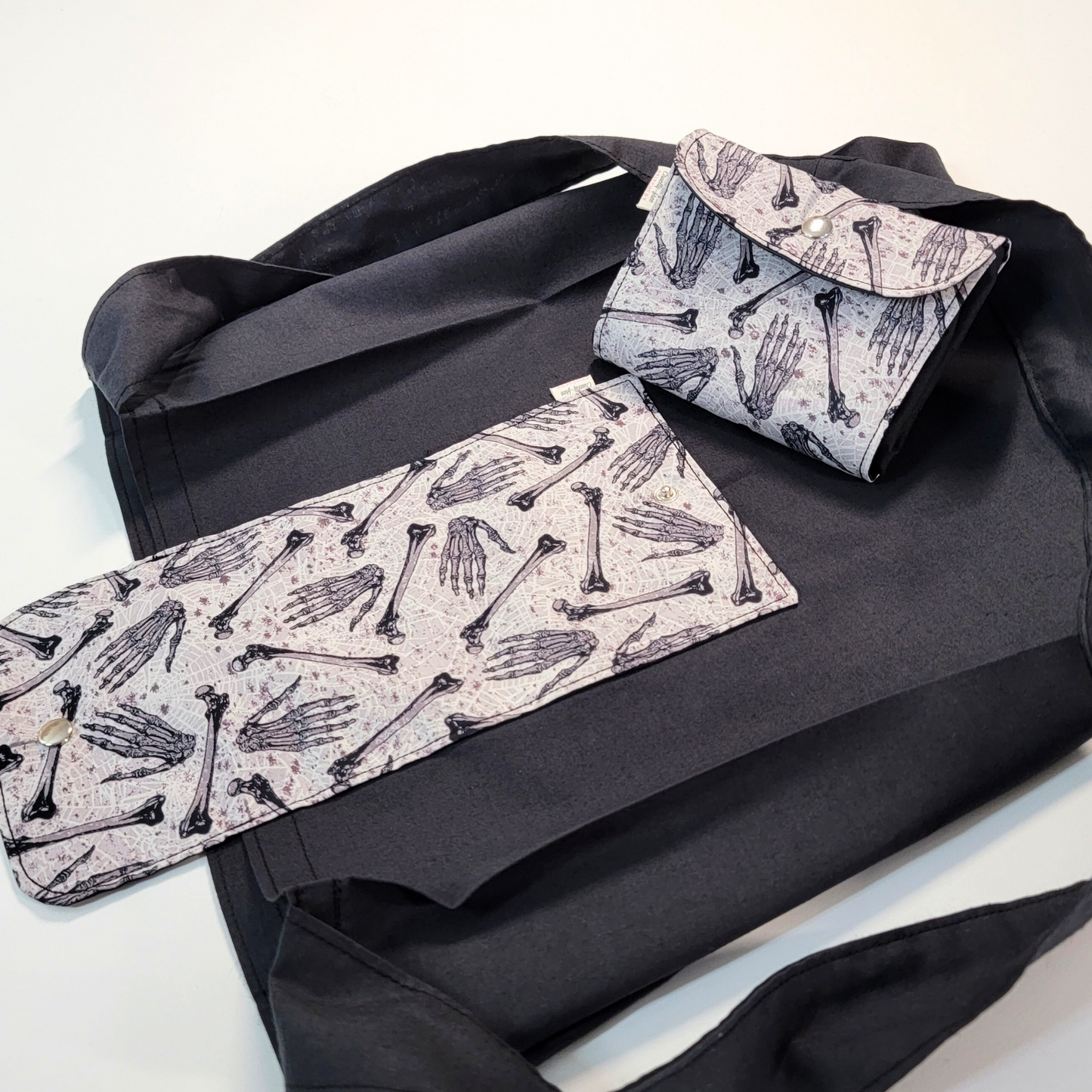 A solid black tote bag with a long strap and a grey flap printed with skeleton hands and femur bones. On top of the bag is another bag that has been folded up and snapped together like a wallet.
