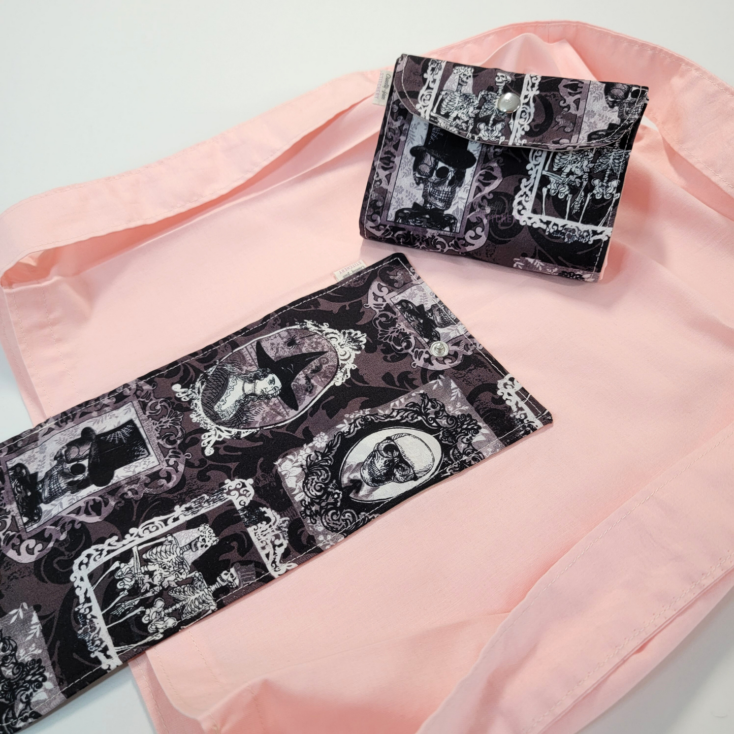A solid light pink tote bag with a long strap and a black and gray flap printed with gothic style portrait frames with skulls, a witch, and a skeleton couple. On top of the bag is another bag that has been folded up and snapped together like a wallet.