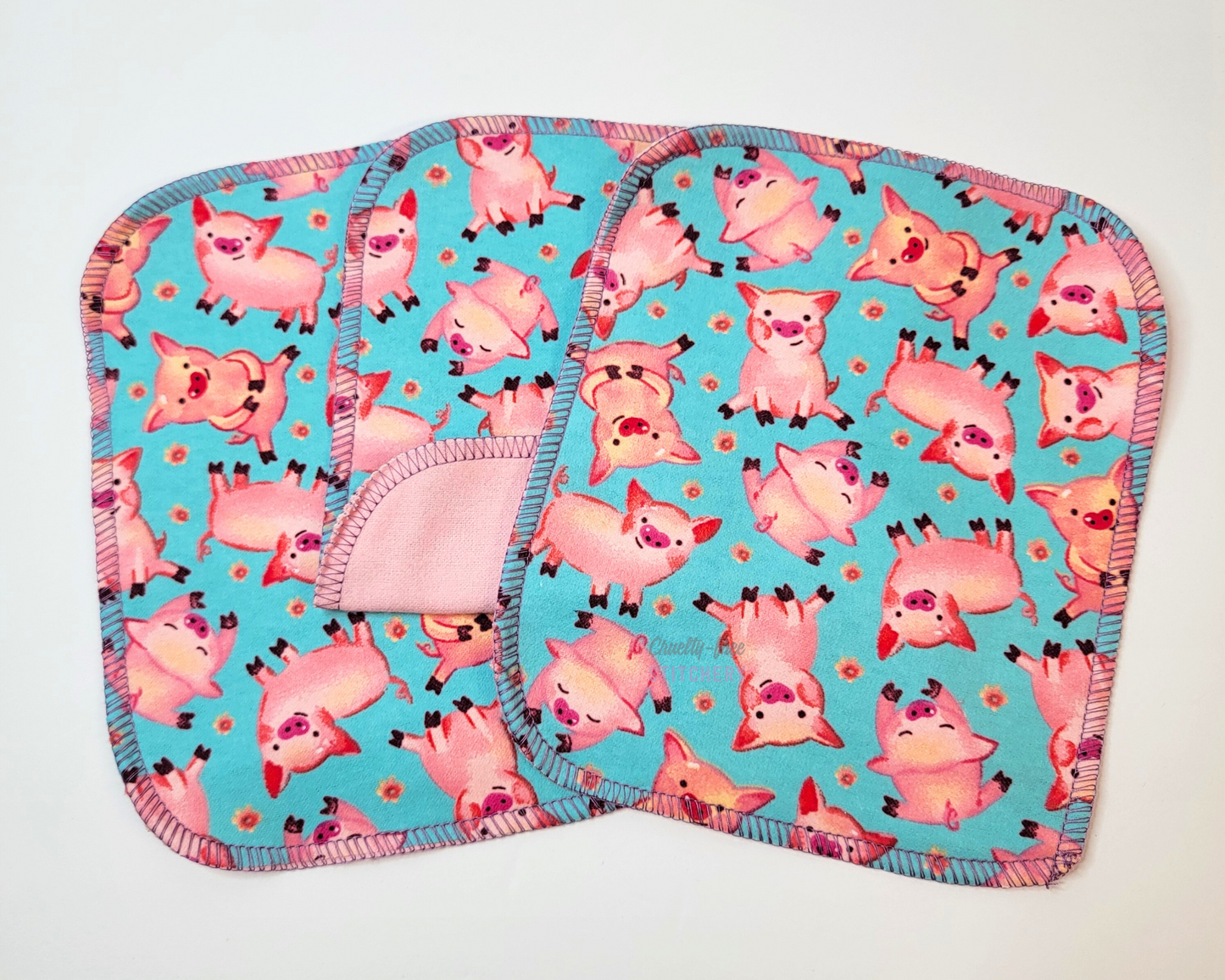 Blue Pigs Reusable Cloth Wipes. Three wipes laid out, with the center one folded back to show the opposite side is light pink. They are a bright aqua blue with small pink pigs and tiny pink flowers.