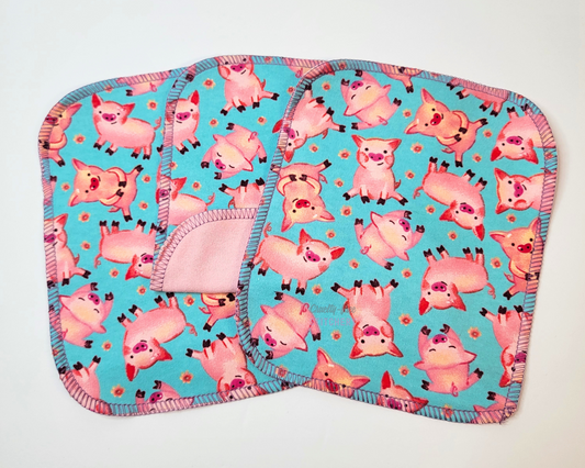 Blue Pigs Reusable Cloth Wipes. Three wipes laid out, with the center one folded back to show the opposite side is light pink. They are a bright aqua blue with small pink pigs and tiny pink flowers. 