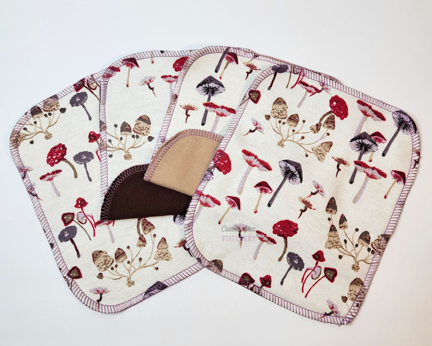 Four mushroom print cloth wipes laid out in a fan shape, the two in the center are folded up to show the back is tan or dark brown. The mushroom print is a white background with wild mushrooms in shades of reds, browns, and purples.