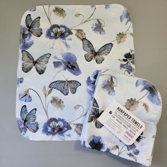 Butterfly Violet print NonPaper Towel laid flat to show the detail of the print, with a bundled 10-pack sitting partially on top of it. The NonPaper Towel is a rectangular shape, with rounded corners, The edges are stitched in off-white thread. The bundle's tag says "NonPaper Towels, the simplicity of a rag with the convenience of a paper towel. Use, wash, roll, reuse. 100% cotton fabric, 100% cellulose thread. Plant based, biodegradable."