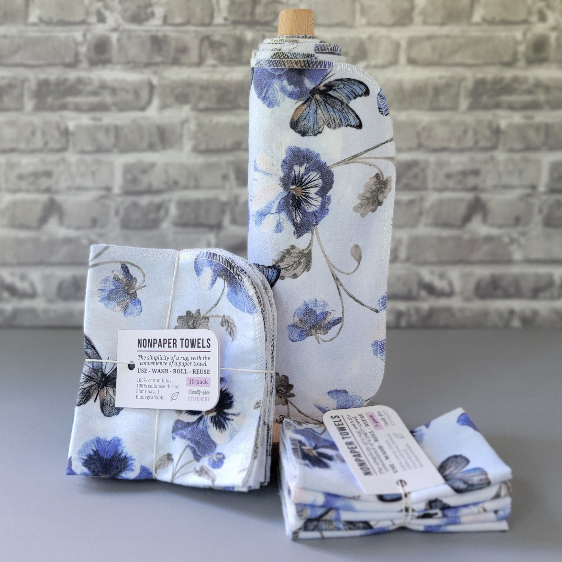 Butterfly Violet print NonPaper Towels. The material is a cloudy pale blue with elegant blue butterflies and blue violets with long gray stems. In the center, they are rolled onto a wooden paper towel holder, and there are bundled packs of 10 and 5 sitting next to the holder. The bundles are tied with string and have a small paper tag. The background is a gray brick wall and solid gray tabletop.