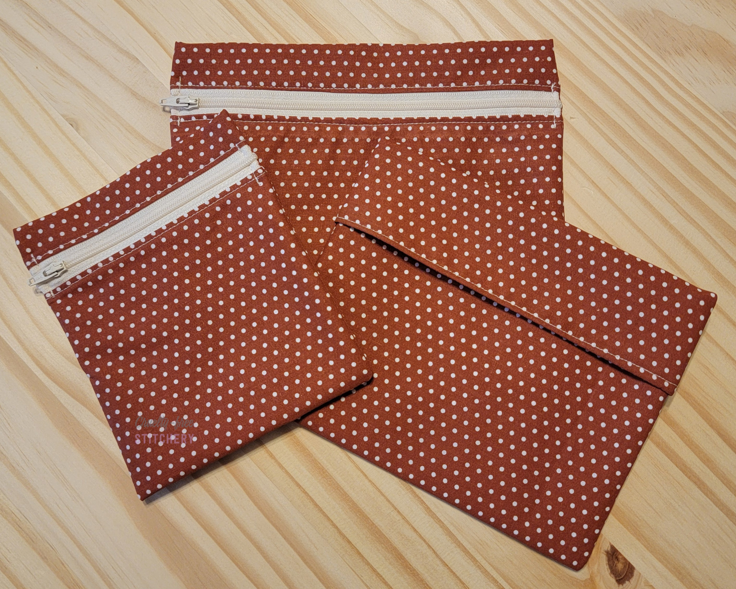 Three sizes of reusable snack bags in the dark orange rust with tiny white dots print. Two sizes have zippers, which are an off-white color.