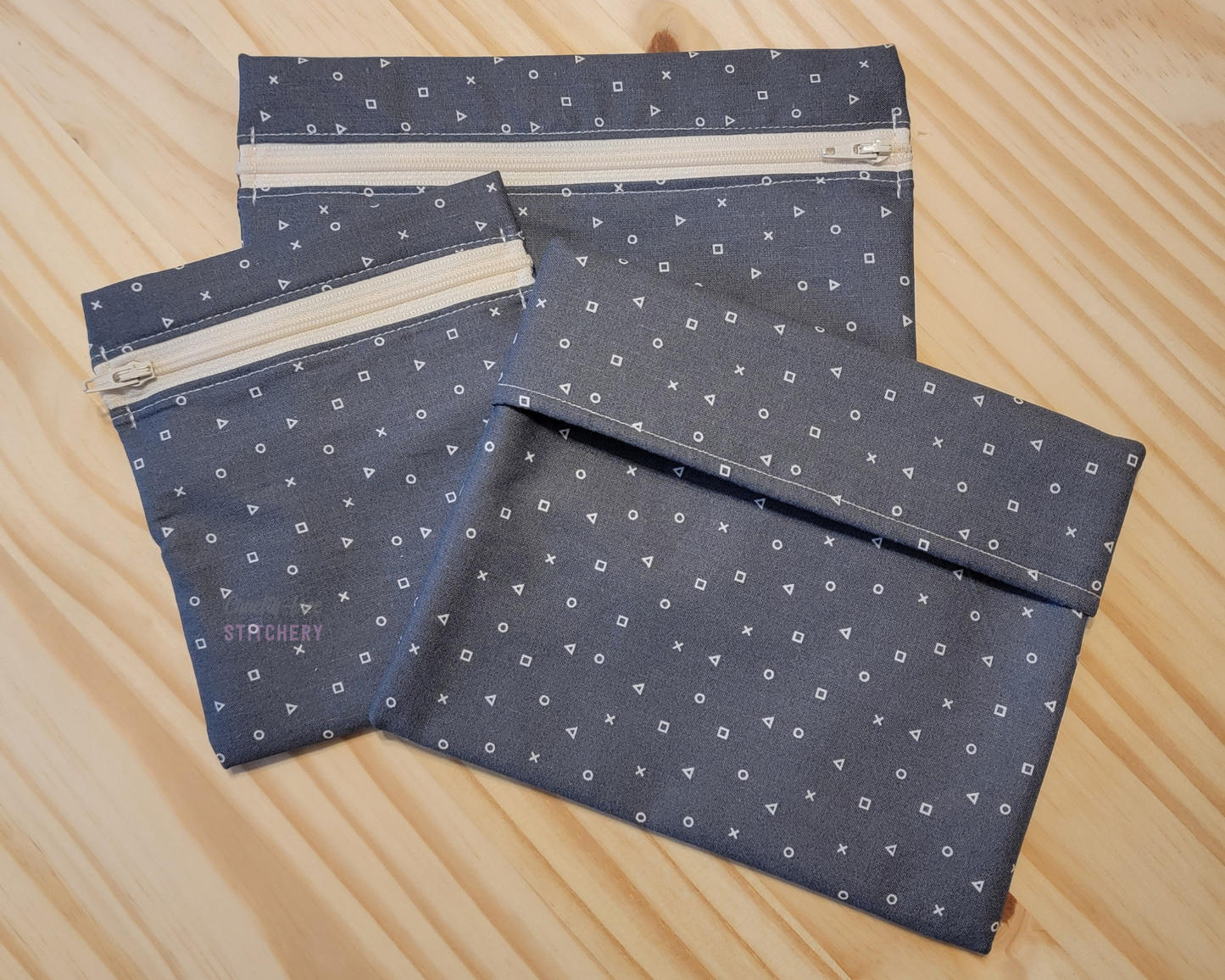 Three sizes of reusable snack bags in the gray with game shapes print. It is a medium-dark gray with tiny x, o, triangle, and square shapes. Two sizes have zippers, which are an off-white color.