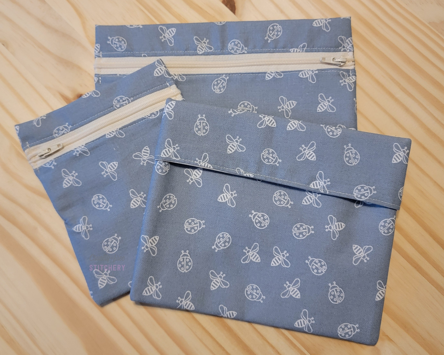 Three sizes of reusable snack bags in the light blue with bugs print. It is a light denim blue with white sketched bees and ladybugs. Two sizes have zippers, which are an off-white color.