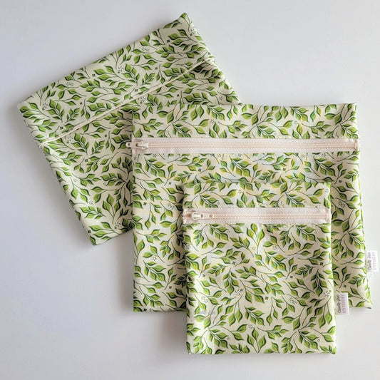 Set of three reusable snack bags with a light olive green leaves print, shown with the foldover style bag closed and behind the zipper styles. The print is a pale green with darker green leaves on stems.