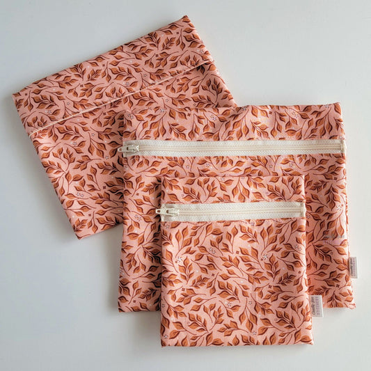 Set of three reusable snack bags with a pink leaves print, shown with the foldover style bag closed and behing the zipper styles. The print is a warmer toned pink background with darker yellowish pink leaves and stems.