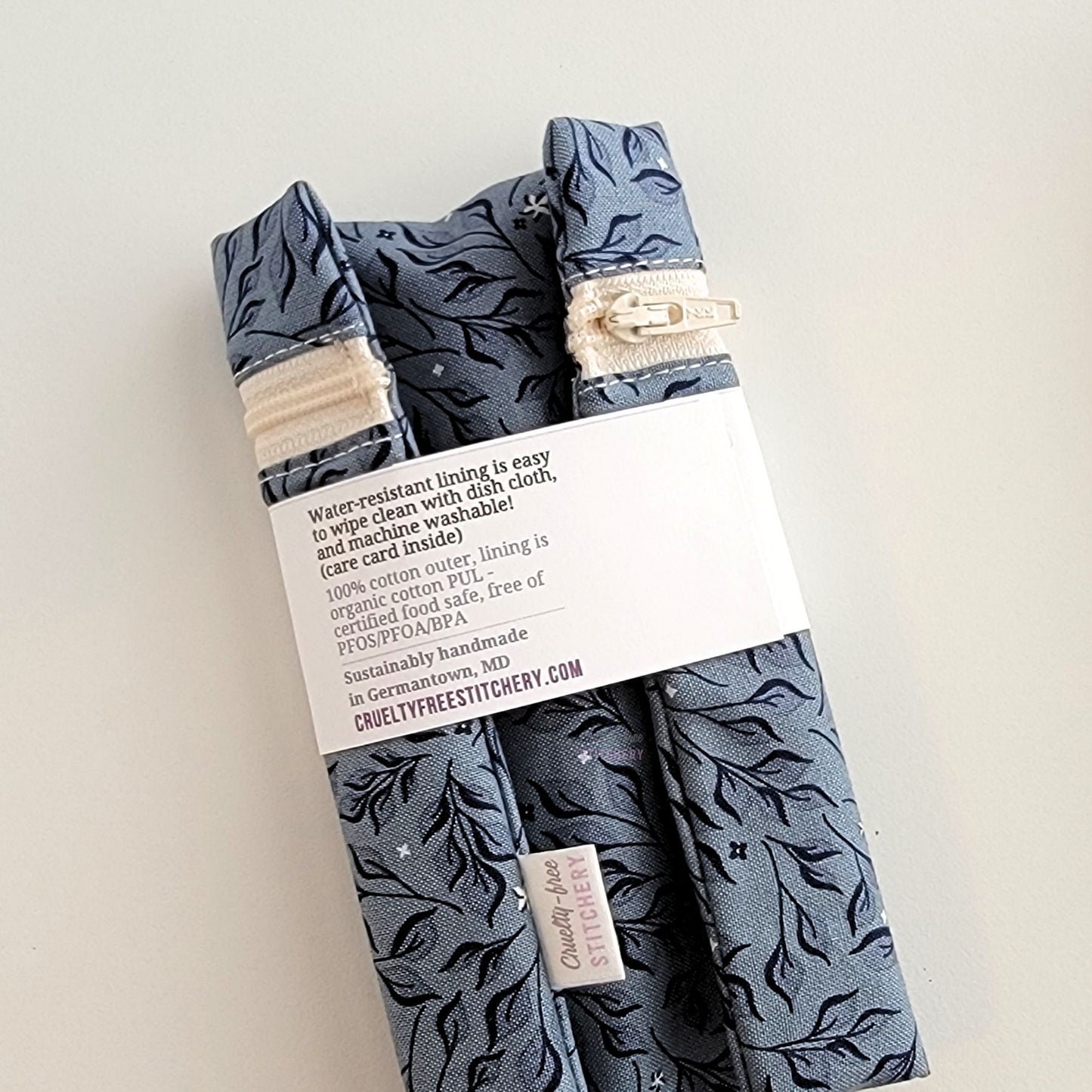 The back of the Stone Leaves Snack Bag packaging. It says "Water resistant lining is easy to wipe clean with dish cloth, and machine washable. Care card inside. 100% cotton outer, lining is organic cotton PUL, cerfitied food-safe, free of PFOS, PFOA, BPA. Sustainable handmade in Germantown, MD" and our website url.