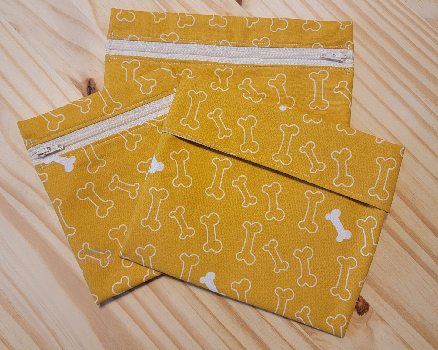 Three sizes of reusable snack bags in the yellow with bones print. It is a mustard yellow with white outlined dog bones, some are filled in. Two sizes have zippers, which are an off-white color.