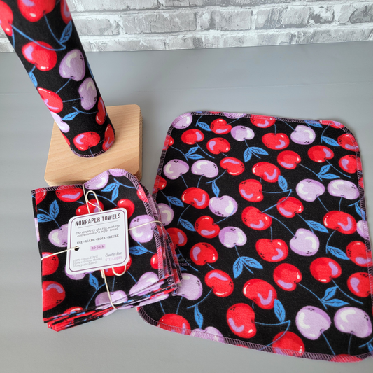 The black cherries nonPaper Towels - one is laid flat to show the print, the cherries are on stems and are approximately an inch wide. The towels have rounded corners and are stitched in a lilac purple thread. Next to that is a bundled pack that is tied with string, and some towels on a wooden holder.