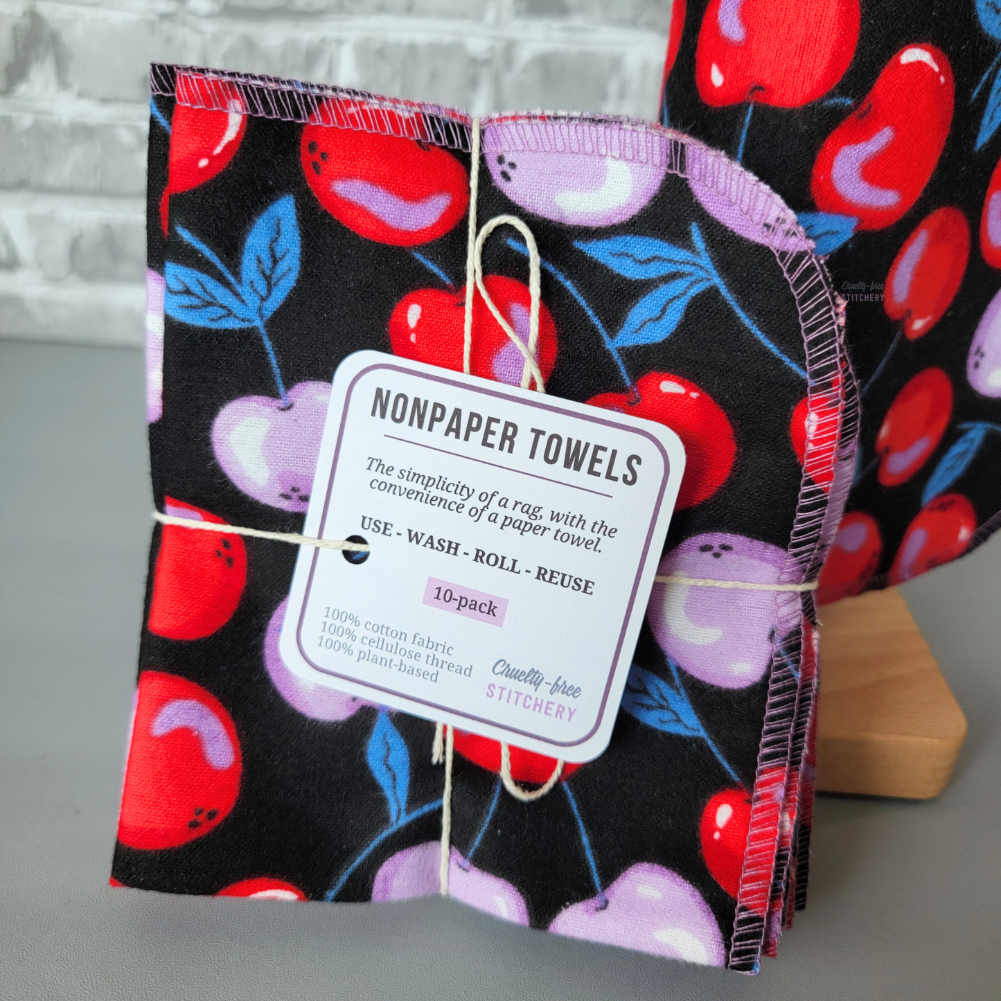 a close-up of the tag on the black cherries NonPaper Towels, it says "nonpaper towels, the simplicity of a rag with the convenience of a paper towel. use wash roll reuse. 10-pack, 100% cotton fabric, cellulose thread, plant-based, Cruelty-Free Stitchery"