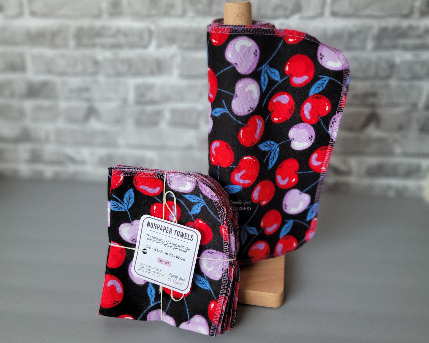 A set of black non-paper towels with a bright red and light purple cherries print, displayed on a wooden stand with a label showing the product name and size.