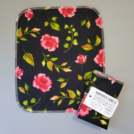 Black with Pink Roses print NonPaper Towel laid flat to show the detail of the print, with a bundled 5-pack sitting partially on top of it. The NonPaper Towel is a rectangular shape, with rounded corners, The edges are stitched in off-white thread. The bundle's tag says "NonPaper Towels, the simplicity of a rag with the convenience of a paper towel. Use, wash, roll, reuse. 100% cotton fabric, 100% cellulose thread. Plant based, biodegradable."