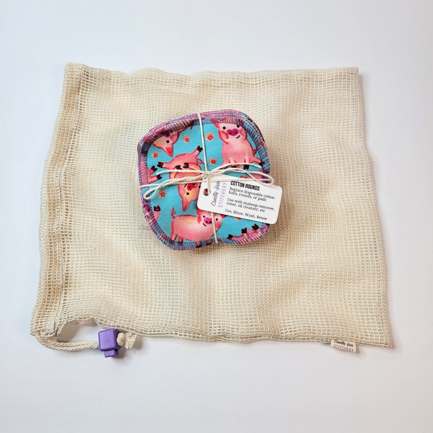 Bundled pack of cotton rounds on top of a cotton mesh drawstring bag. The bag is a natural off-white colored square mesh, with a light purple cord stop.