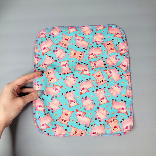 A blue pigs print NonPaper Towel shown flat on a table with my hand for size reference and print detail. The background is bright aqua blue, with tossed pink pigs in different poses, some sitting, standing, or laying down, with tiny pink flowers in-between. The towel measures approximately 10 by 12 inches, and there are about 7 pigs across the width and 8 across the length.