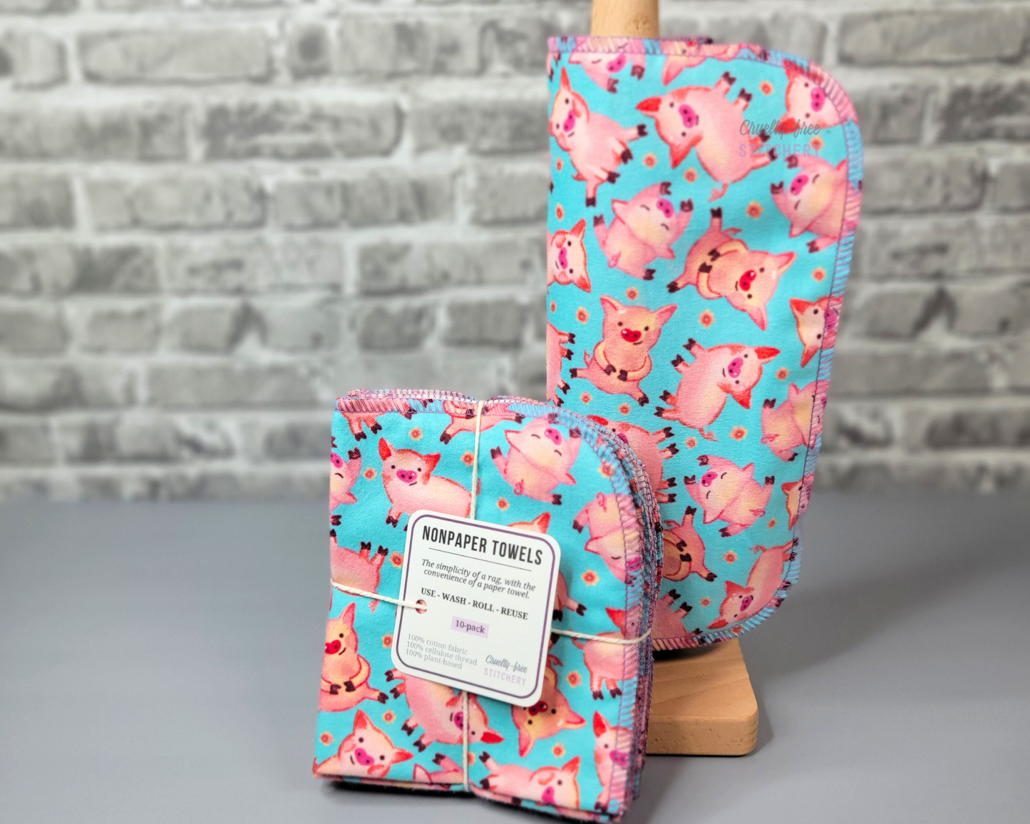 Aqua Blue with pink pigs print NonPaper Towels. They are rolled onto a wooden paper towel holder, and sitting next to a bundled pack of 10. The bundled pack is folded and tied with a string, the tag says "nonpaper towels, use, wash, roll, reuse, the simplicity of a rag with the convenience of a paper towel." The edges of each NonPaper Towel are stitched with a lilac purple thread, with the corners rounded.