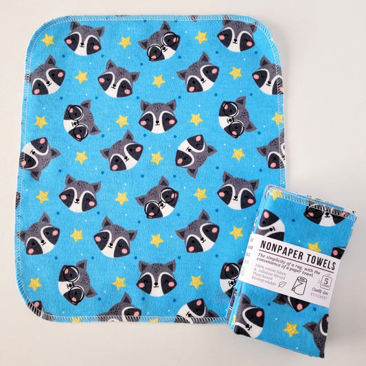 An unfolded Blue Raccoons NonPaper Towel, next to a bundled 5-pack. The towel is a rectangle with rounded corners, stitched around the edges with white thread. The bundled pack is folded up with a paper wrapper that says "NonPaper Towels, the simplicity of a rag with the convenience of a paper towel"