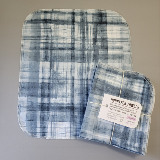 Blue Watercolor Plaid print NonPaper Towel laid flat to show the detail of the print, with a bundled 10-pack sitting partially on top of it. The NonPaper Towel is a rectangular shape, with rounded corners, The edges are stitched in off-white thread. The bundle's tag says "NonPaper Towels, the simplicity of a rag with the convenience of a paper towel. Use, wash, roll, reuse. 100% cotton fabric, 100% cellulose thread. Plant based, biodegradable."
