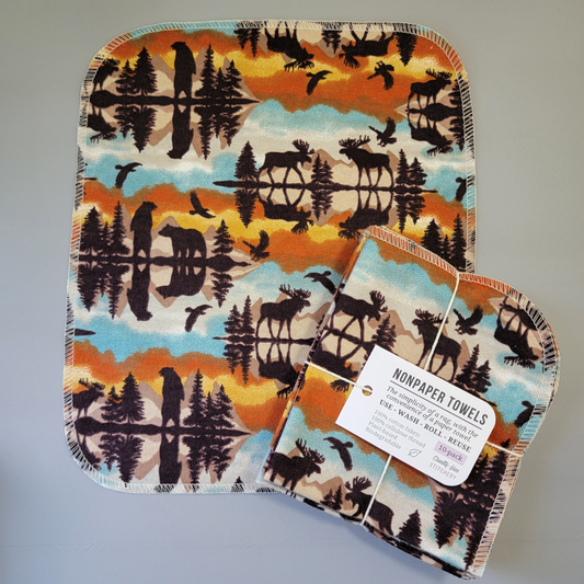 Cabin View print NonPaper Towel laid flat to show the detail of the print, with a bundled 10-pack sitting partially on top of it. The NonPaper Towel is a rectangular shape, with rounded corners, The edges are stitched in off-white thread. The bundle's tag says "NonPaper Towels, the simplicity of a rag with the convenience of a paper towel. Use, wash, roll, reuse. 100% cotton fabric, 100% cellulose thread. Plant based, biodegradable."