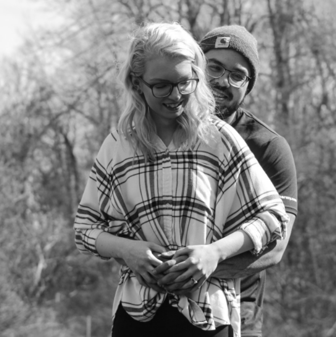 Black and white photo of Abby in a white plaid shirt, smiling and being hugged from behind by her husband - wearing a beanie and short sleeved shirt.