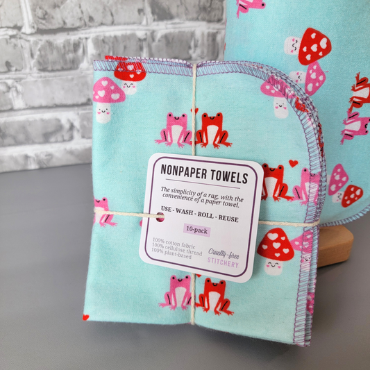 A stack of 10 light aqua blue non-paper towels with a pink and red frog and mushroom pattern. The towels are tied with twine and have a label that reads 'NONPAPER TOWELS' 'The simplicity of a rag, with the convenience of a paper towel.' 'USE-WASH-ROLL-REUSE' '10-pack' '100% cotton fabric' '100% cellulose thread' '100% plant-based' 'Cruelty-free STITCHERY'.