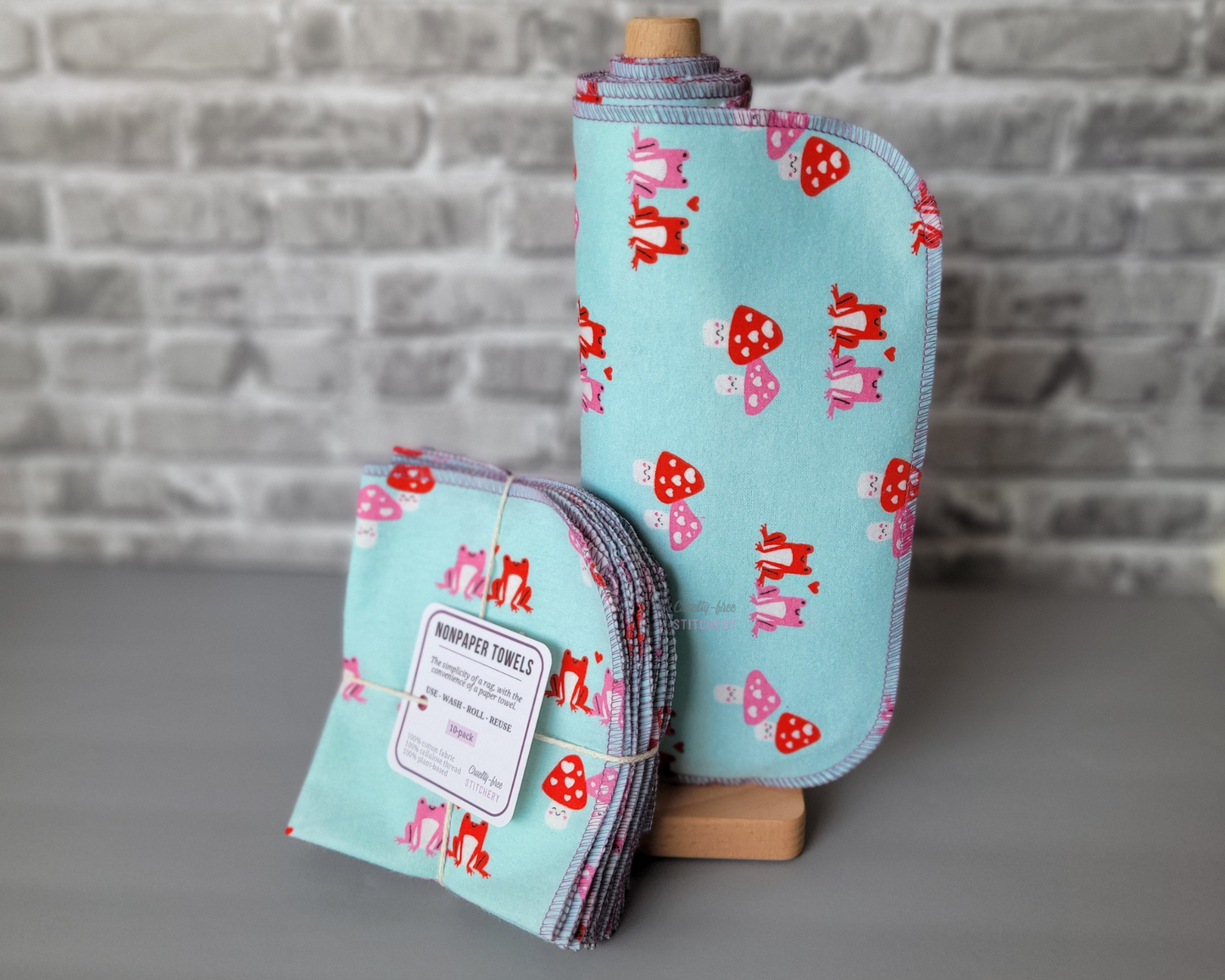 A set of NonPaper Towels - light aqua blue with small pink and red frogs and pink and red mushrooms with faces in a Valentine's day style. Some are rolled onto a wooden holder, next to a bundled pack tied with string and a small paper tag.