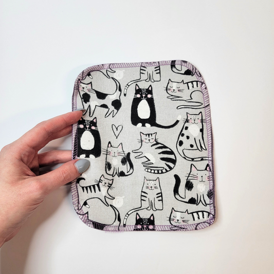 May include: A light gray fabric square with a purple border. The fabric has a repeating pattern of black and white cats in various poses. Some of the cats have hearts above their heads.