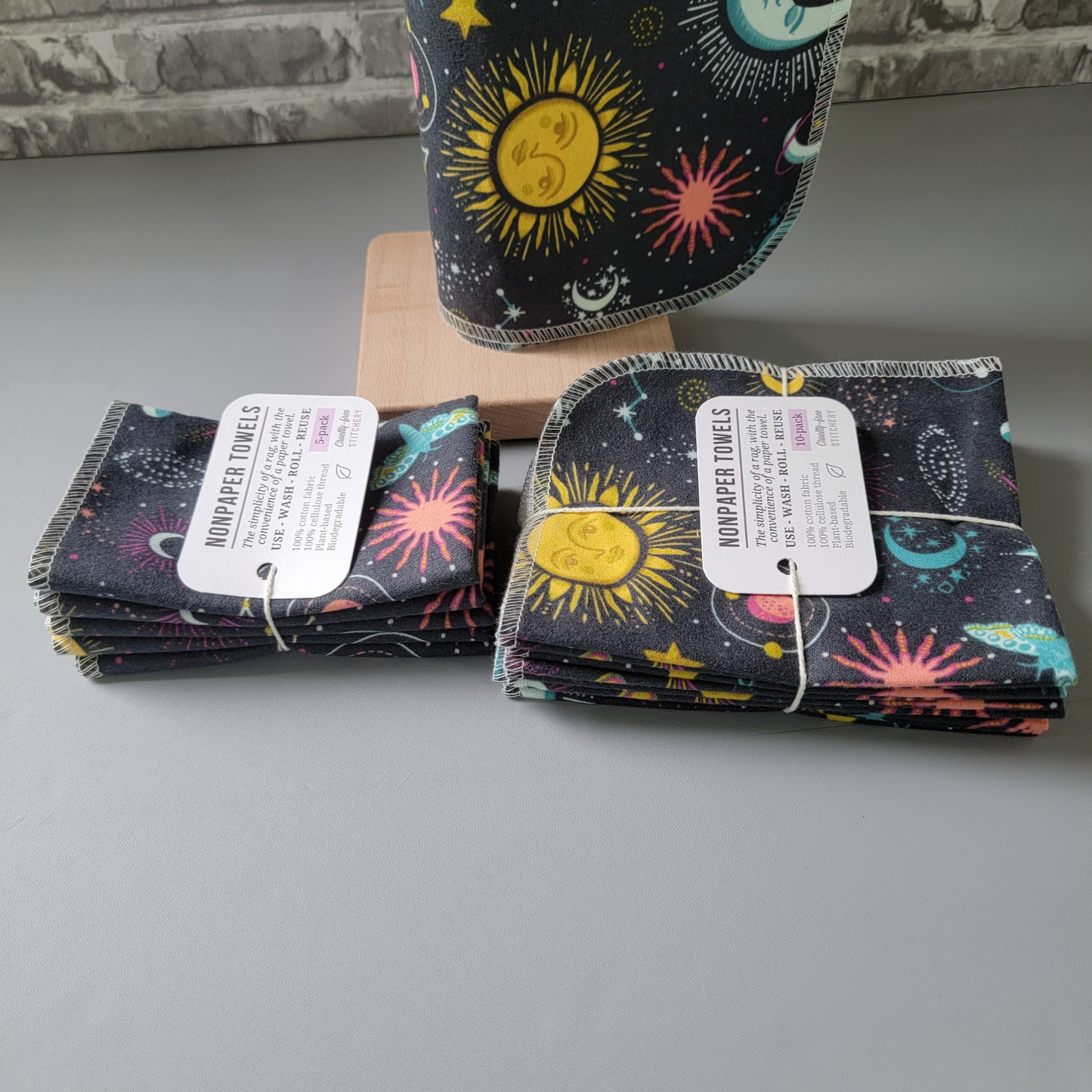A 5-pack and 10-pack laid next to each other, photographed from the side to show the difference. The 10-pack has the towels folded in half twice, while the 5-pack has them folded one more time so they look smaller. Both are tied with cotton string and a white paper tag.