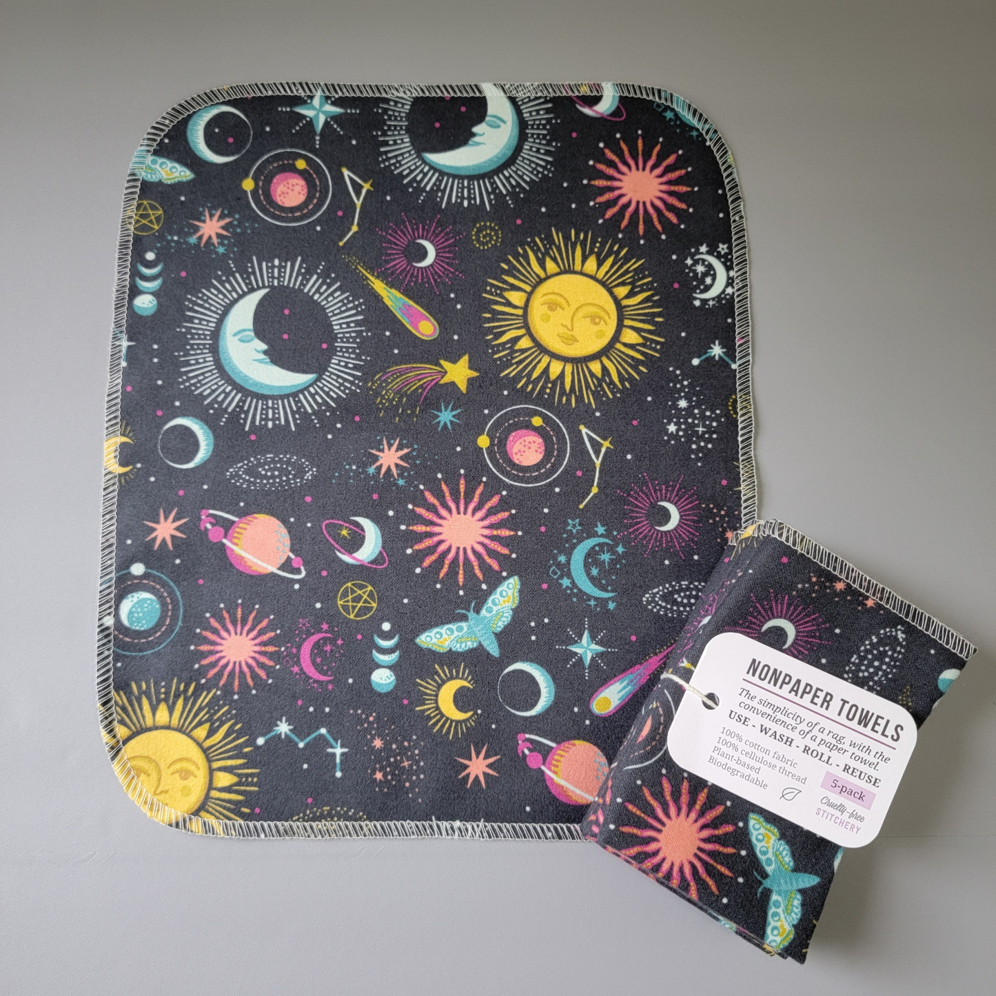 A gray celestial nonpaper towel laid flat to show the print better, next to it is a bundled 5-pack. Lots of different cosmic icons, it's like an i-spy picture.