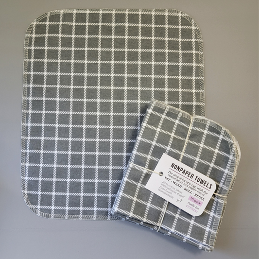 A Gray Small Windowpane print NonPaper Towel laid flat to show the detail of the print, with a bundled 10-pack sitting partially on top of it. The NonPaper Towel is a rectangular shape, with rounded corners, The edges are stitched in off-white thread. The bundle's tag says "NonPaper Towels, the simplicity of a rag with the convenience of a paper towel. Use, wash, roll, reuse. 100% cotton fabric, 100% cellulose thread. Plant based, biodegradable."