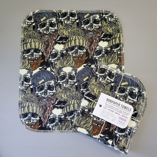 A Hipster Skulls print NonPaper Towel laid flat to show the detail of the print, with a bundled pack of 10 sitting partially on top of it. The NonPaper Towel is a rectangular shape, with rounded corners, The edges are stitched in off-white thread. The bundle's tag says "NonPaper Towels, the simplicity of a rag with the convenience of a paper towel. Use, wash, roll, reuse. 100% cotton fabric, 100% cellulose thread. Plant based, biodegradable."