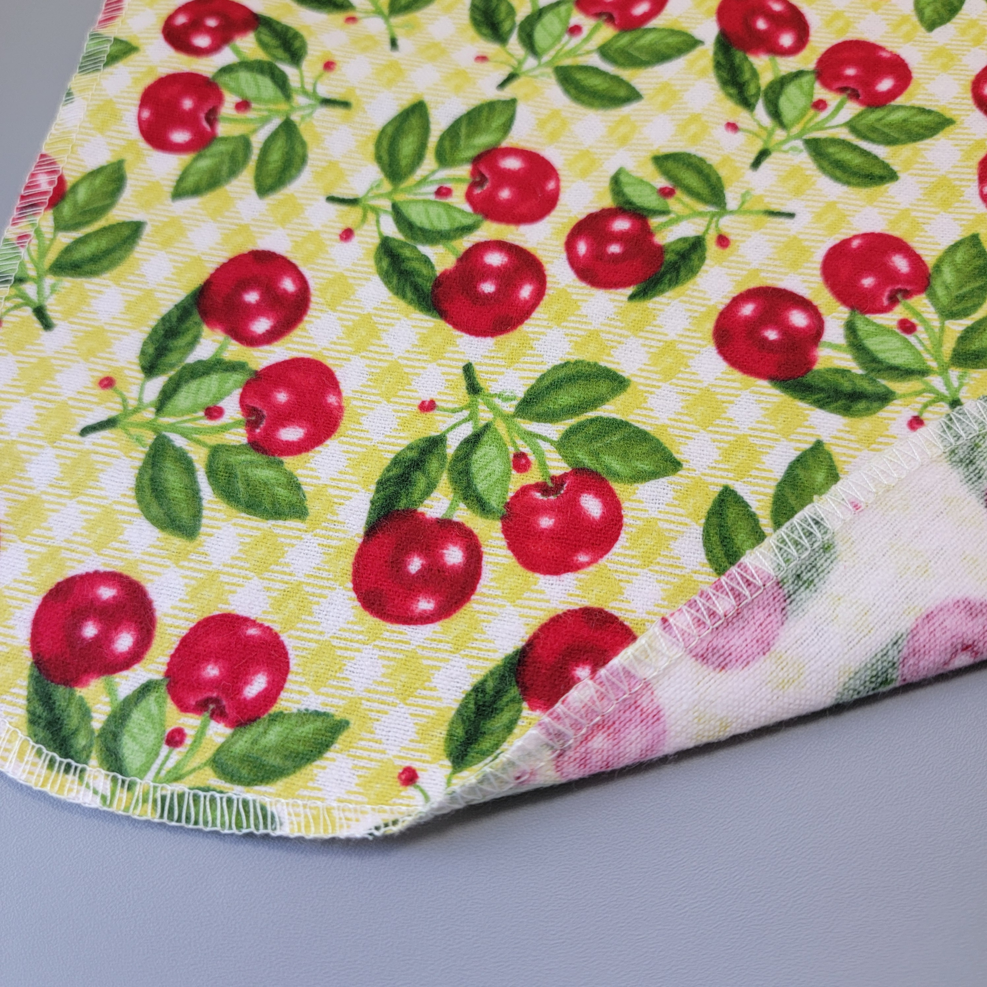 The bottom edge of a Kitschy Cherries NonPaper Towel close-up to show the detail of the stitching. The edge is partially folded back to show the back side is unprinted, and there is one layer of material.