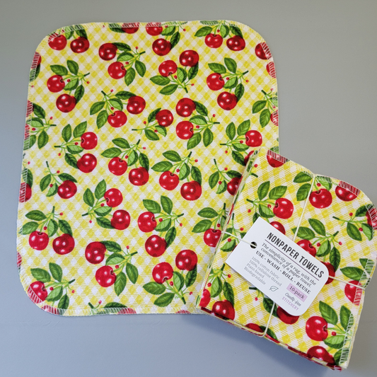 Kitschy Cherries print NonPaper Towel laid flat to show the detail of the print, with a bundled 10-pack sitting partially on top of it. The NonPaper Towel is a rectangular shape, with rounded corners, The edges are stitched in off-white thread. The bundle's tag says "NonPaper Towels, the simplicity of a rag with the convenience of a paper towel. Use, wash, roll, reuse. 100% cotton fabric, 100% cellulose thread. Plant based, biodegradable."