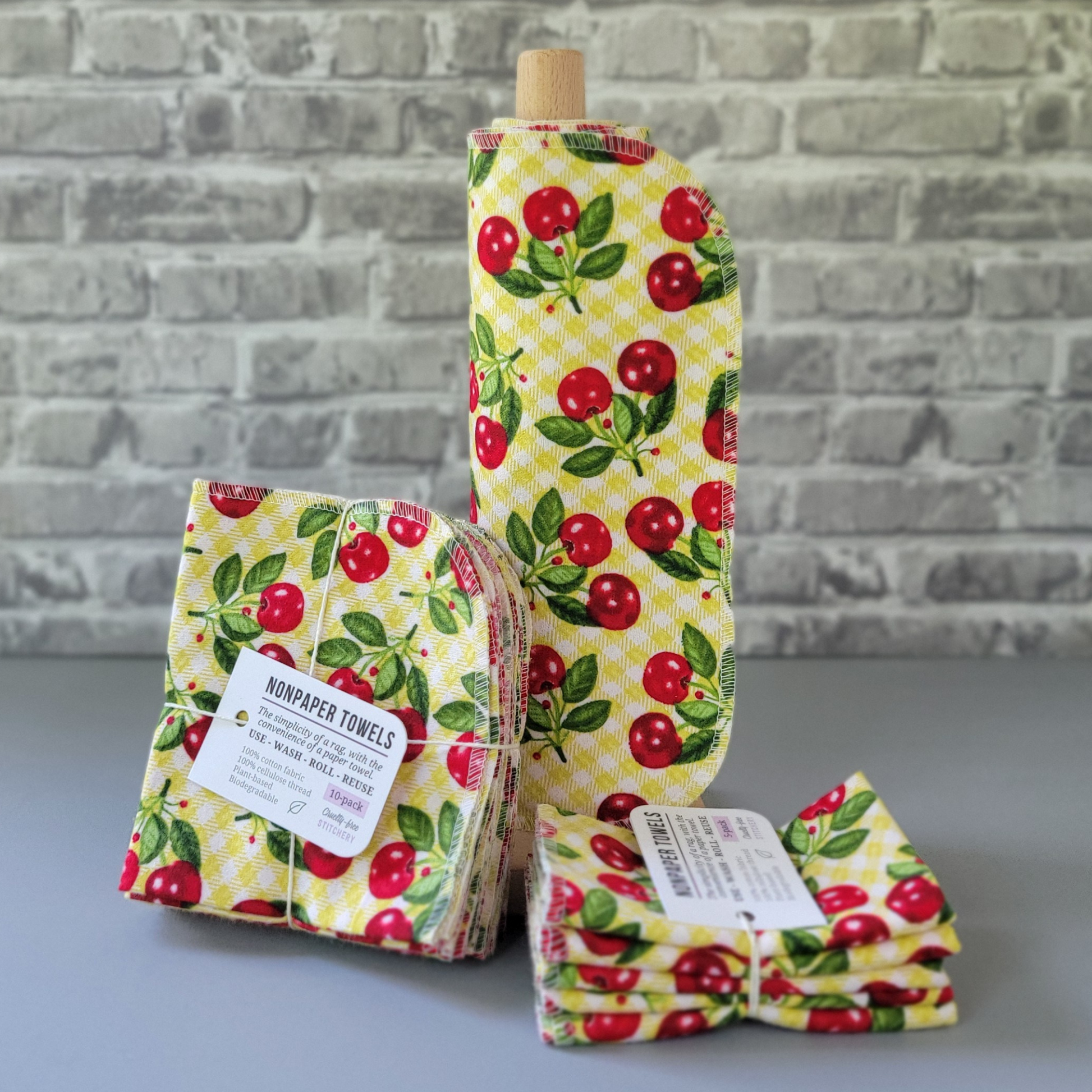 Kitschy Cherries print NonPaper Towels. The print is vintage-style cherries in pairs with the stems, on an obnoxious chartreuse or yellow-green and white gingham background. In the center, they are rolled onto a wooden paper towel holder, and there are bundled packs of 10 and 5 sitting next to the holder. The bundles are tied with string and have a small paper tag. The background is a gray brick wall and solid gray tabletop.