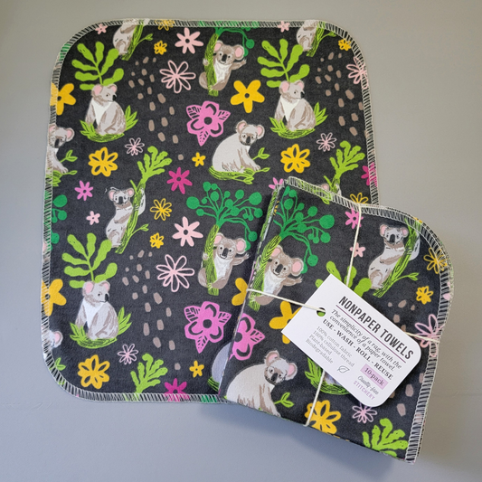 Koalas print NonPaper Towel laid flat to show the detail of the print, with a bundled 10-pack sitting partially on top of it. The NonPaper Towel is a rectangular shape, with rounded corners, The edges are stitched in off-white thread. The bundle's tag says "NonPaper Towels, the simplicity of a rag with the convenience of a paper towel. Use, wash, roll, reuse. 100% cotton fabric, 100% cellulose thread. Plant based, biodegradable."