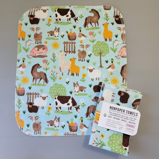 Light Blue Farm print NonPaper Towel laid flat to show the detail of the print, with a bundled 5-pack sitting partially on top of it. The NonPaper Towel is a rectangular shape, with rounded corners, The edges are stitched in off-white thread. The bundle's tag says "NonPaper Towels, the simplicity of a rag with the convenience of a paper towel. Use, wash, roll, reuse. 100% cotton fabric, 100% cellulose thread. Plant based, biodegradable."