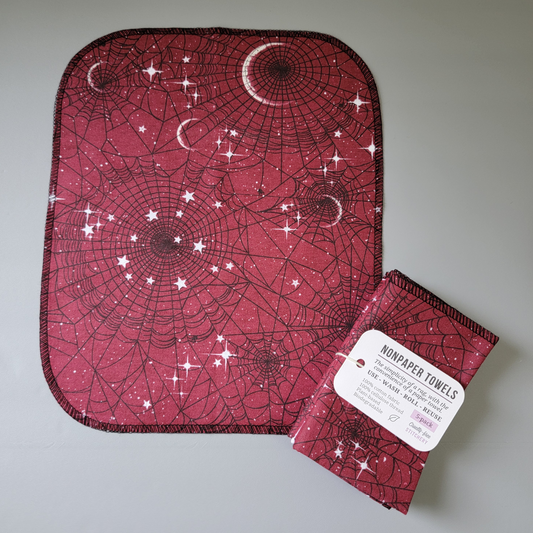 A red moon web NonPaper Towel laid flat to show the pattern. Black spider webs of varying sizes and clusters of stars and moon shapes. The edges are rounded and stitched with black thread. Laying partially on top is a bundled 5-pack, with a tag that says "nonpaper towels, the simplicity of a rag with the convenience of a paper towel. Use wash roll reuse. 100% cotton fabric, 100% cellulose thread, plant-based, biodegradable. 5-pack, cruelty-free stitchery, with a tiny leaf.