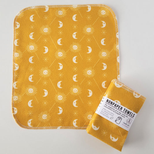 An unfolded Mustard yellow celestial NonPaper Towel, next to a bundled 5-pack. The towel is a rectangle with rounded corners, stitched around the edges with white thread. The bundled pack is folded up with a paper wrapper that says "NonPaper Towels, the simplicity of a rag with the convenience of a paper towel"