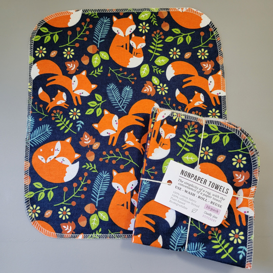 Navy Foxes print NonPaper Towel laid flat to show the detail of the print, with a bundled 10-pack sitting partially on top of it. The NonPaper Towel is a rectangular shape, with rounded corners, The edges are stitched in off-white thread. The bundle's tag says "NonPaper Towels, the simplicity of a rag with the convenience of a paper towel. Use, wash, roll, reuse. 100% cotton fabric, 100% cellulose thread. Plant based, biodegradable."