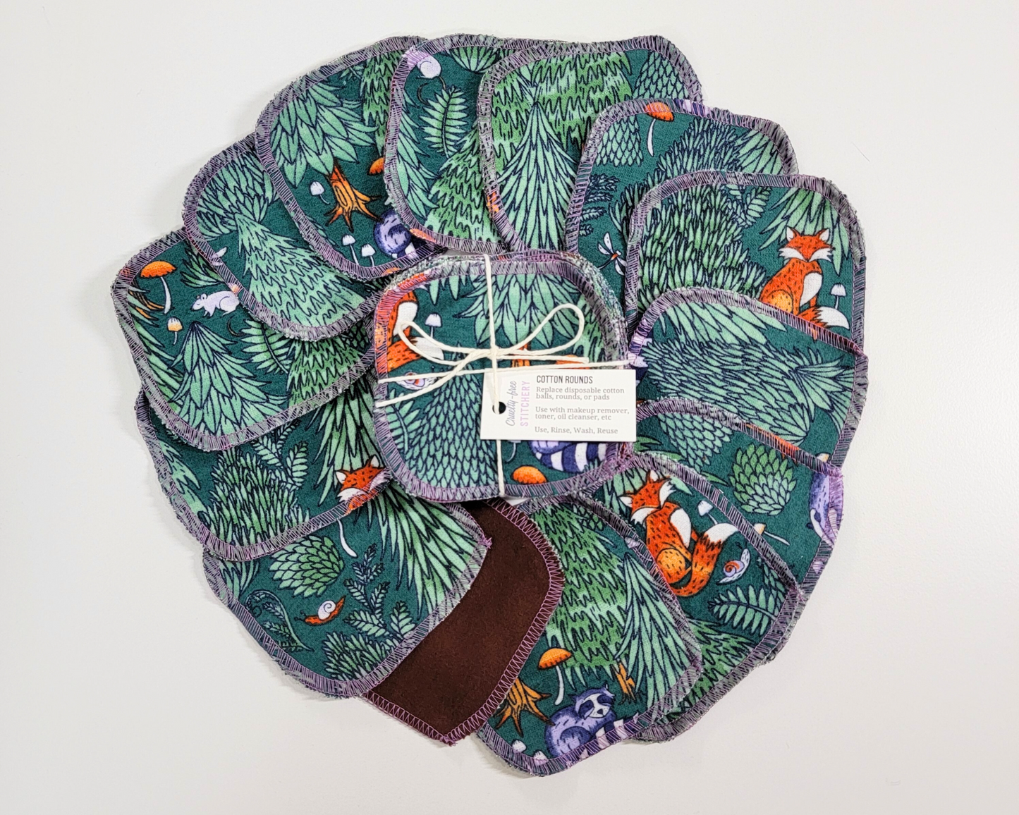 Woodland print reusable cotton rounds arranged in a circle with a bundled pack in the center. They are dark green with trees and animals like foxes, raccoons, mice, etc. One is flipped over to show that the back is dark brown.